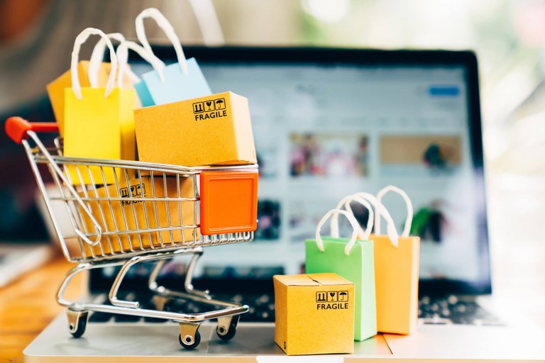 picture of laptop computer in the background, with a miniature shopping cart filled with an assortment of bags inside and outside the cart